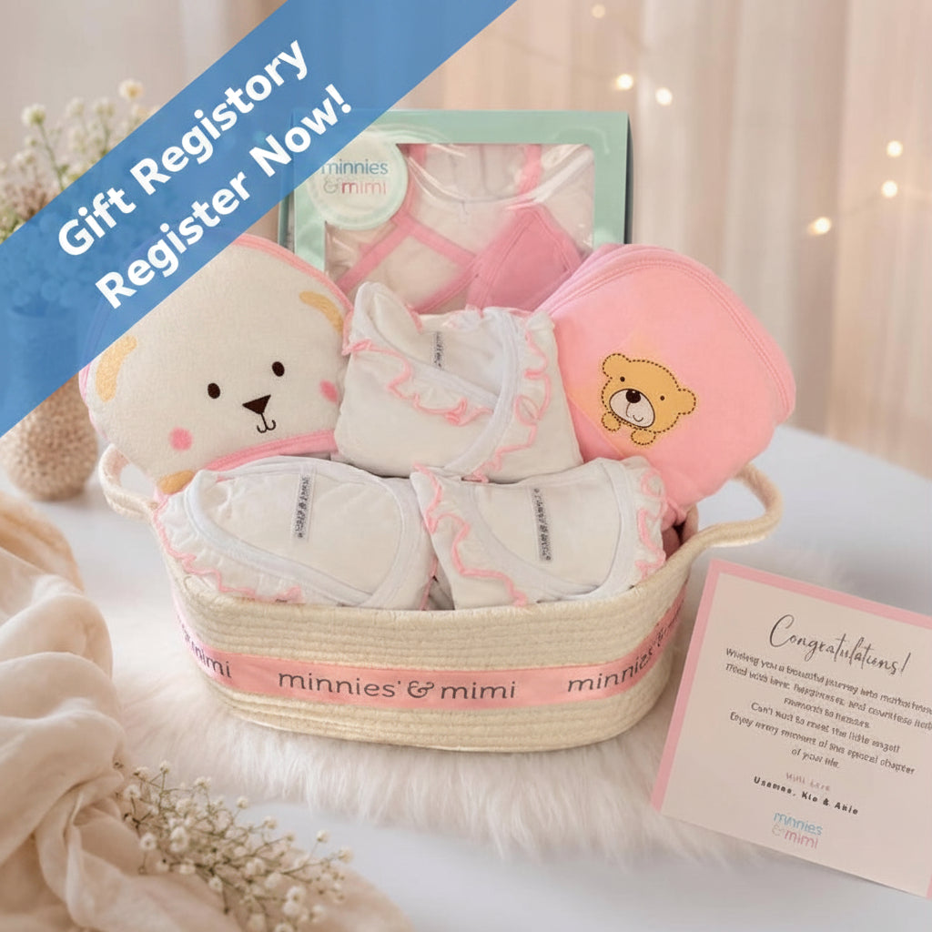 A baby gift basket set from minnies & mimi, featuring a woven straw basket with a pink ribbon, white and pink baby clothes, a plush bear toy, a pink hooded towel, and a congratulations card, styled on a white faux fur surface with soft background bokeh.